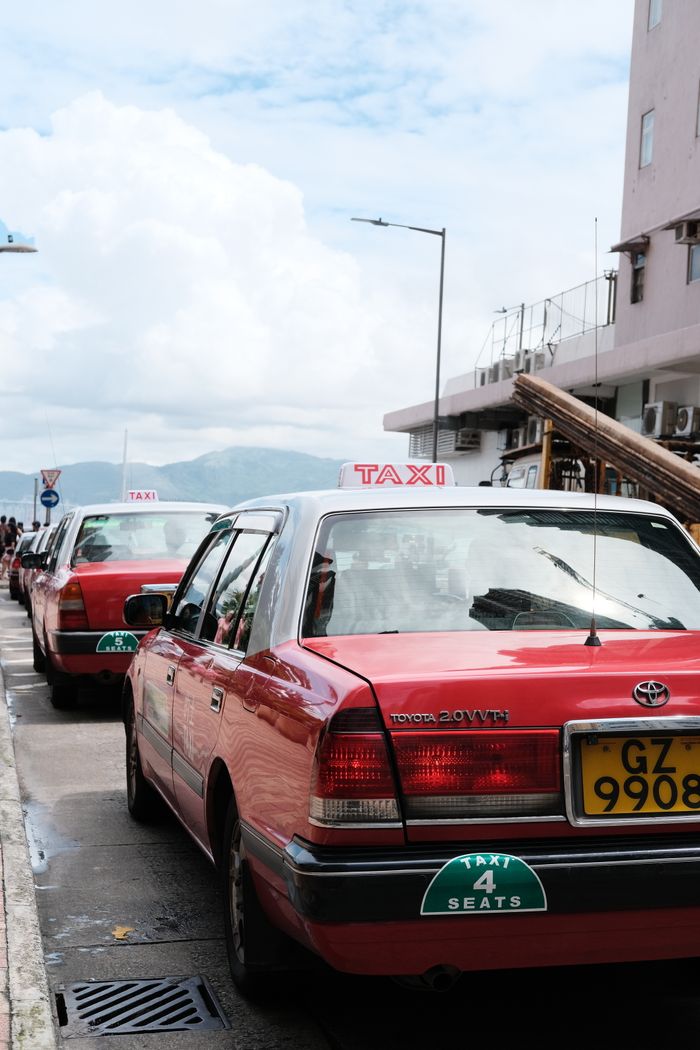 services-img Red taxis parked along a street in a city with a clear sunny sky, ready for passengers.