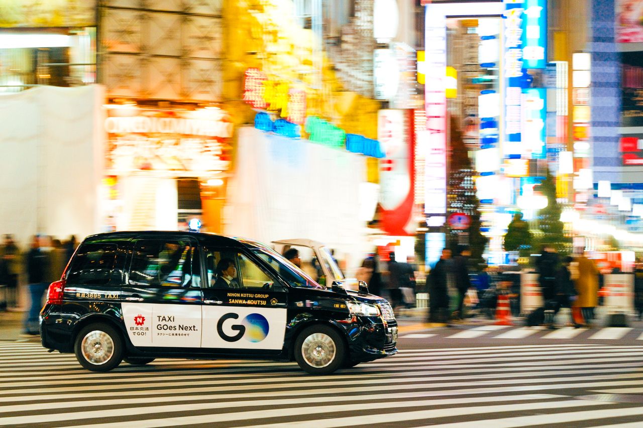 about-img A black taxi driving through a bustling, colorful city street at night under bright neon lights.